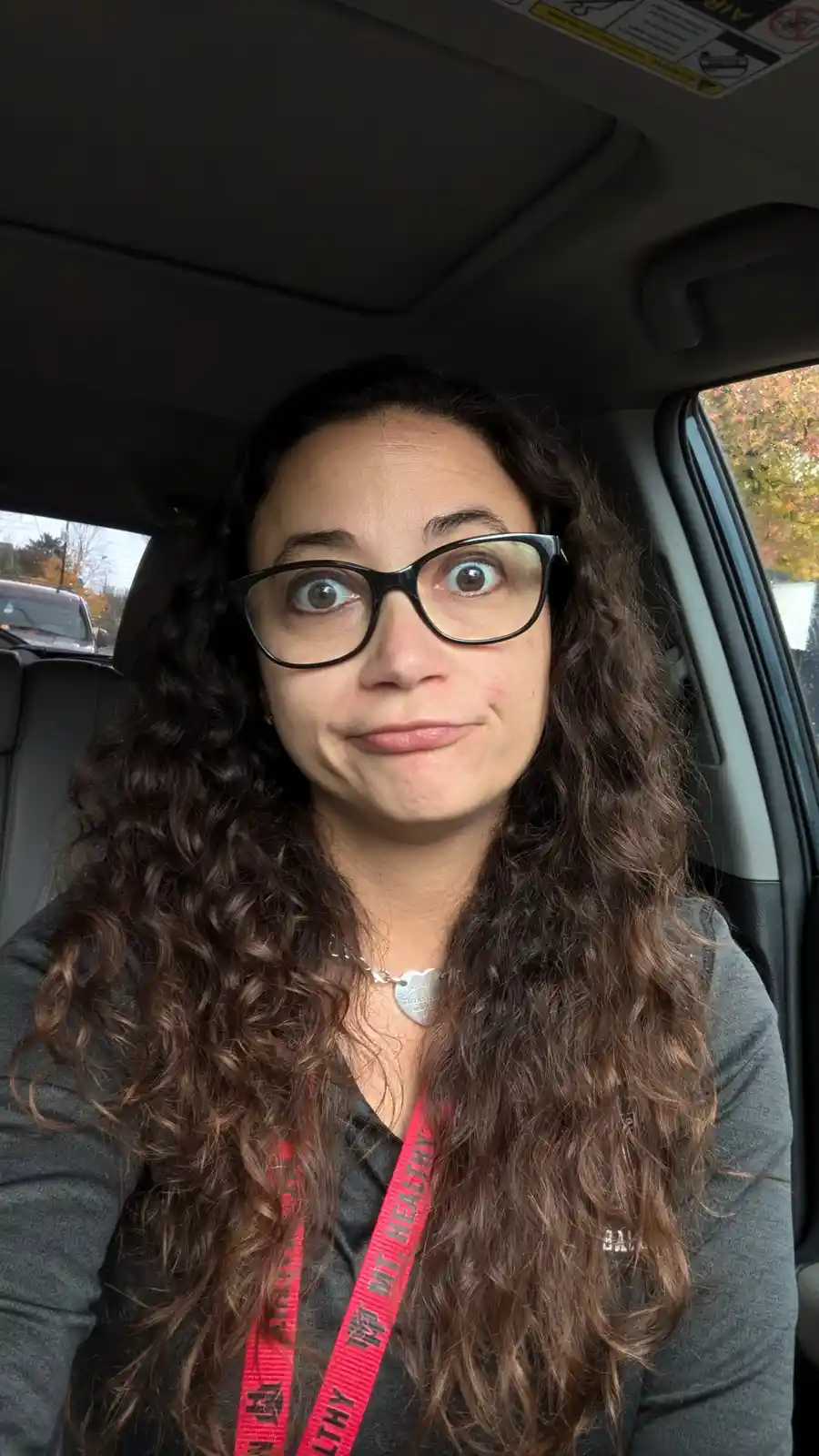 A woman with long curly brown hair and black-rimmed glasses sits in a car, making a wide-eyed, slightly smug facial…