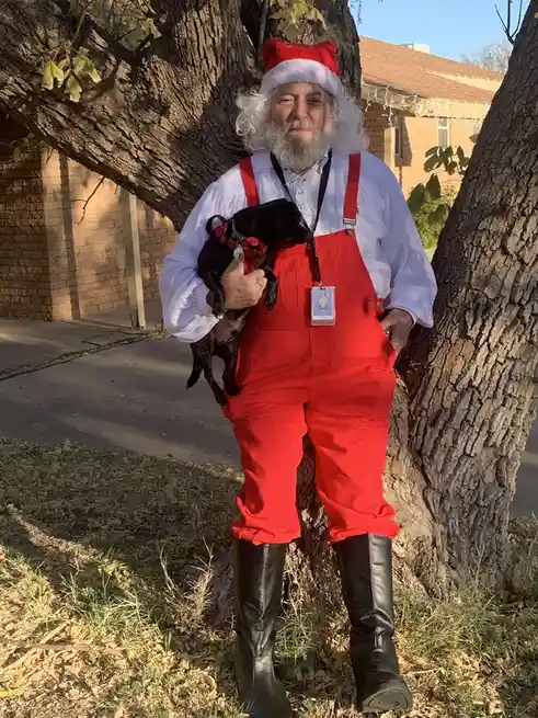 A man dressed as Santa Claus in a red jumpsuit, white beard, and hat, leans against a tree while holding a small black dog…