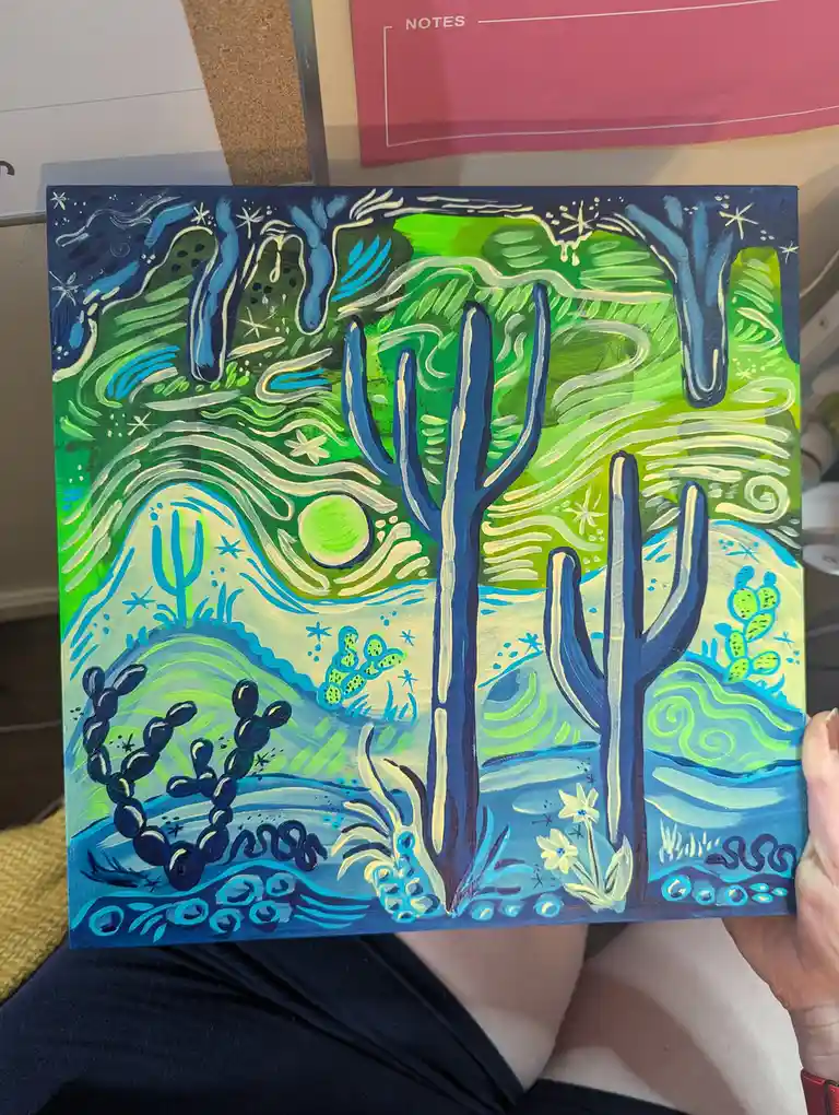 A hand holds up a vibrant, stylized painting depicting a desert scene with cacti and mountains, but rendered in underwater…