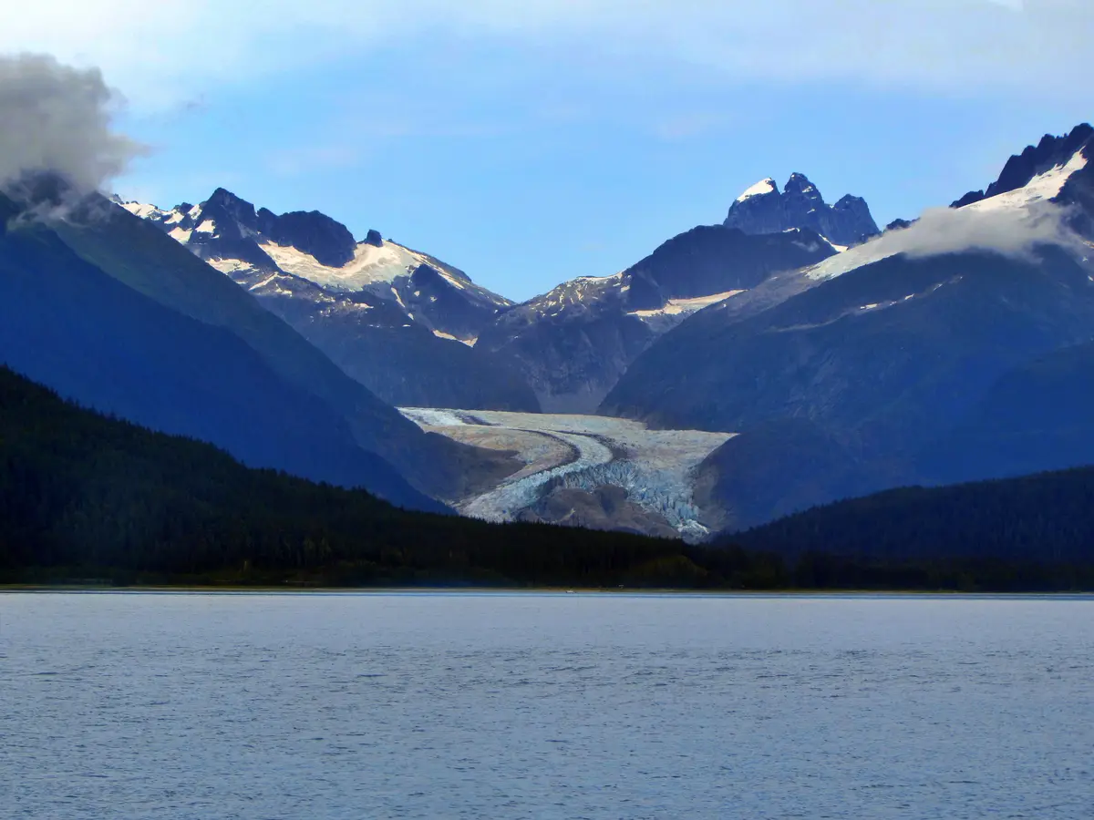 A scenic photograph shows a wide glacier flowing down a valley between snow-capped mountains, with a calm body of water in…