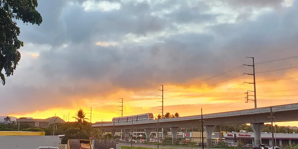 A train travels on an elevated track against a dramatic sunset sky with orange and gray clouds. Below, commercial buildings…