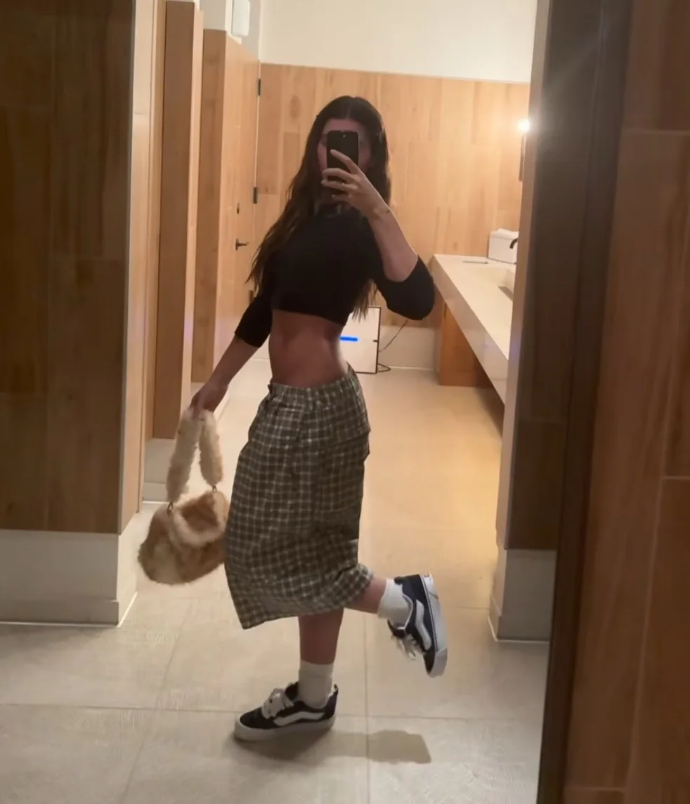 A young woman with long dark hair takes a mirror selfie in a bathroom, wearing a black crop top, a plaid skirt, white socks…