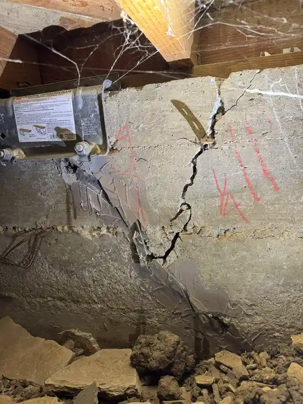 A cracked concrete foundation wall with red spray-painted graffiti and a metal foundation plate bolted to it, surrounded by…