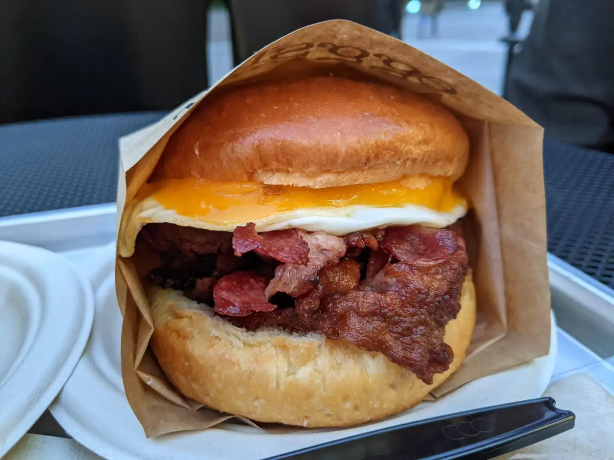 A close-up photo shows a breakfast sandwich in a brown paper wrapper, featuring a toasted bun, a fried egg with runny yolk…