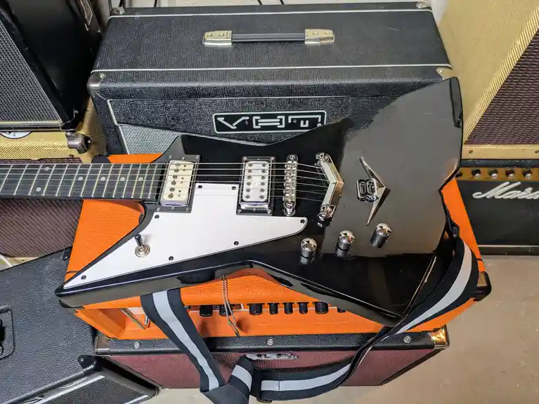 A black electric guitar with a white pickguard and silver hardware rests on top of a stack of orange and black guitar…