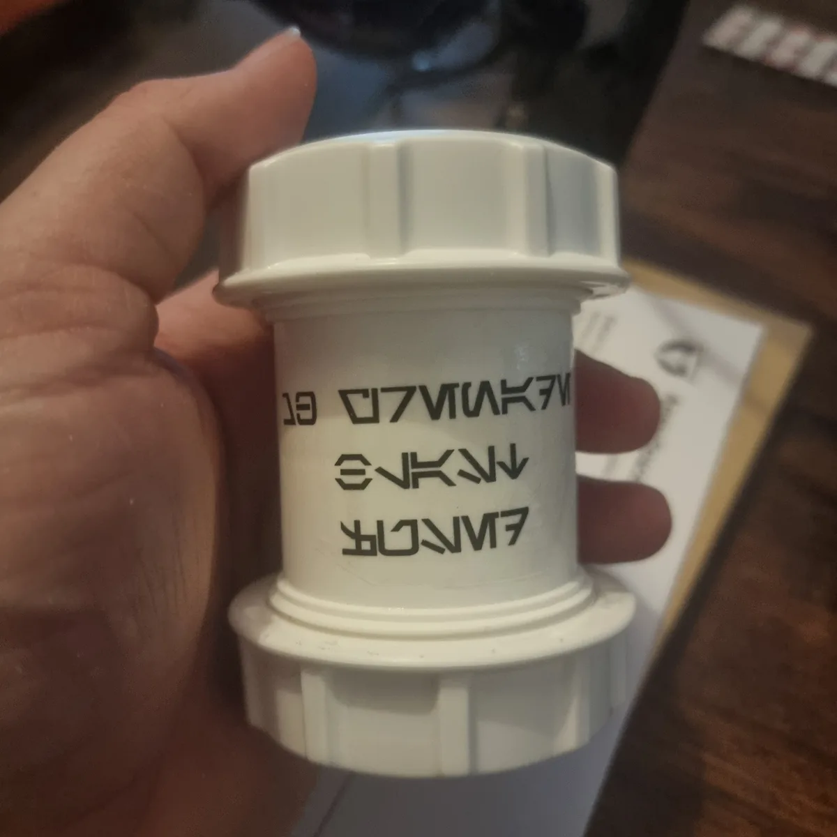 A hand holds a white plastic plumbing fitting with black text that reads 'MAKE ALO EKAN ↓ AMROR', which is gibberish when…