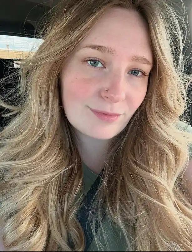 A selfie of a young woman with long blonde hair and blue eyes, smiling slightly while sitting inside a car. She has freckles…