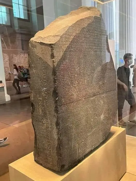 A large, weathered stone slab with ancient inscriptions in three scripts is displayed behind glass in a museum. Visitors are…