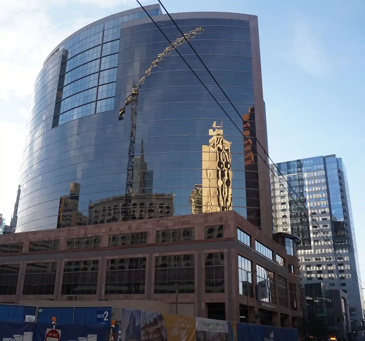 A tall, curved glass skyscraper reflects surrounding buildings and a crane, creating a distorted, fragmented image that…