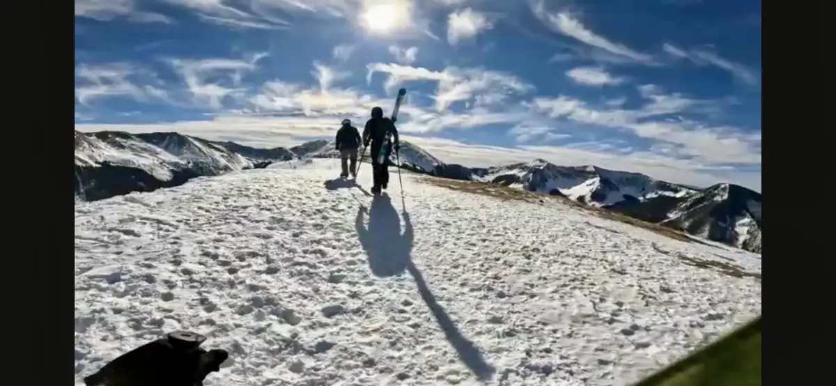 Two people are hiking uphill on a snowy mountain ridge under a bright blue sky with wispy clouds. The person in the…