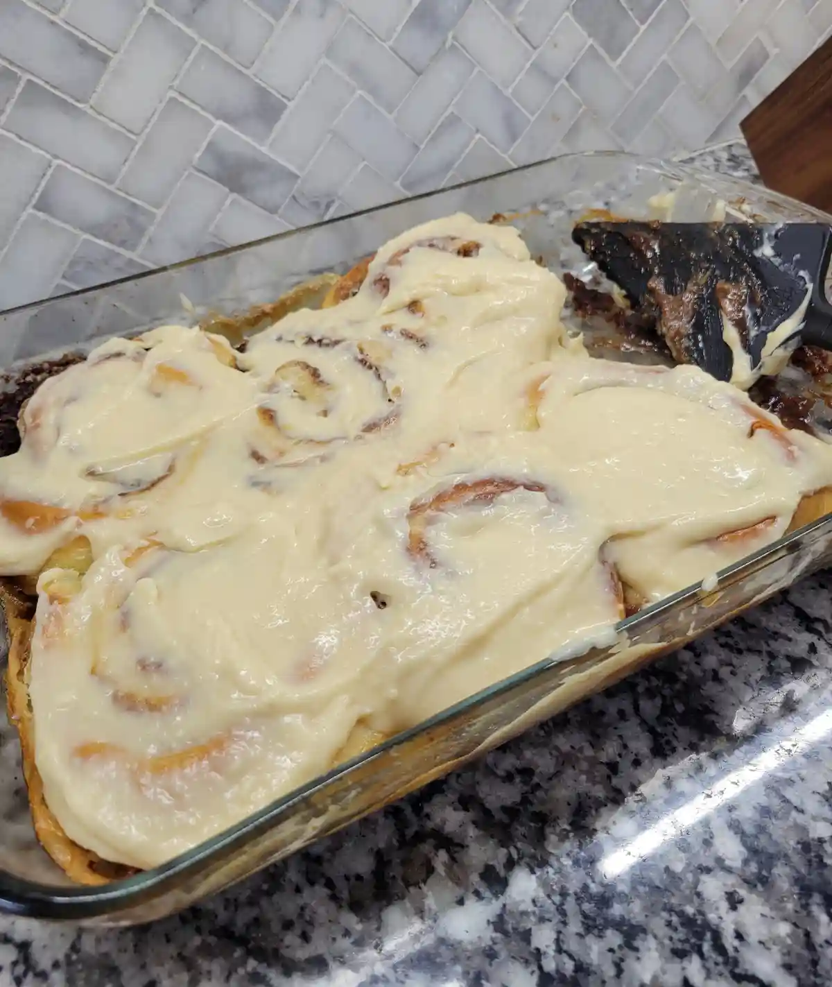 A glass baking dish filled with cinnamon rolls covered in a thick, creamy white frosting, sitting on a granite countertop…