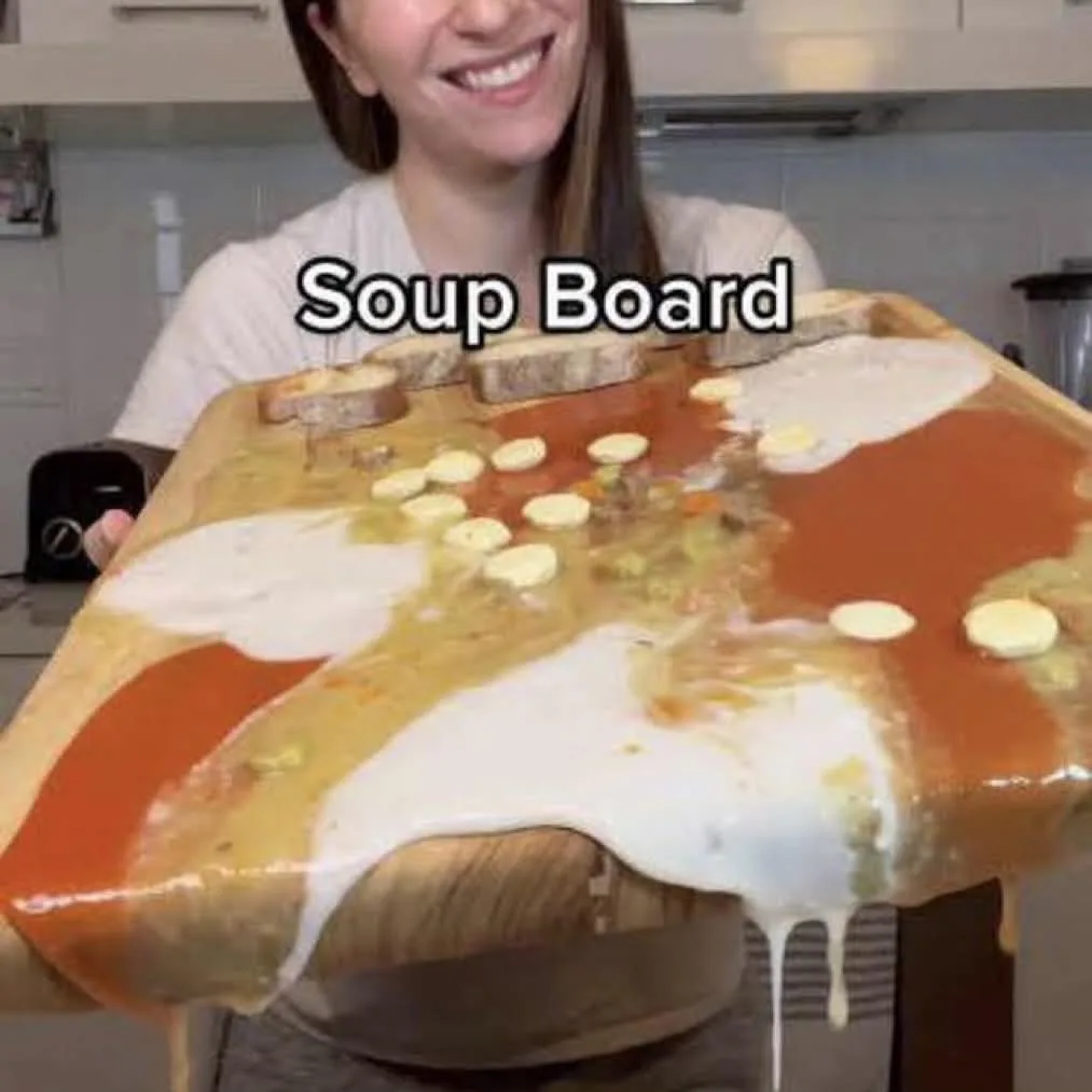 A smiling woman holds up a wooden board covered in a messy, colorful mixture of different soups, with bread slices and small…