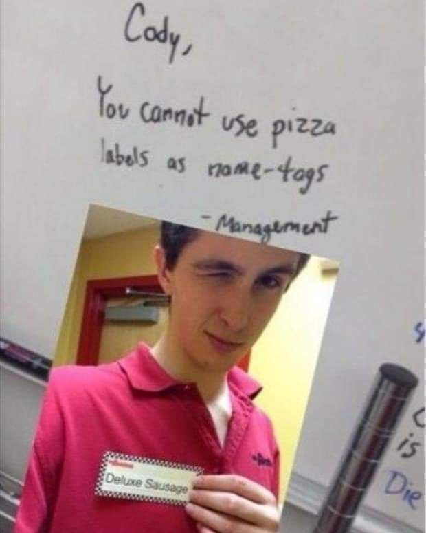 A photo of a young man in a red polo shirt holding a pizza label that reads 'Deluxe Sausage' as a name tag, with a…