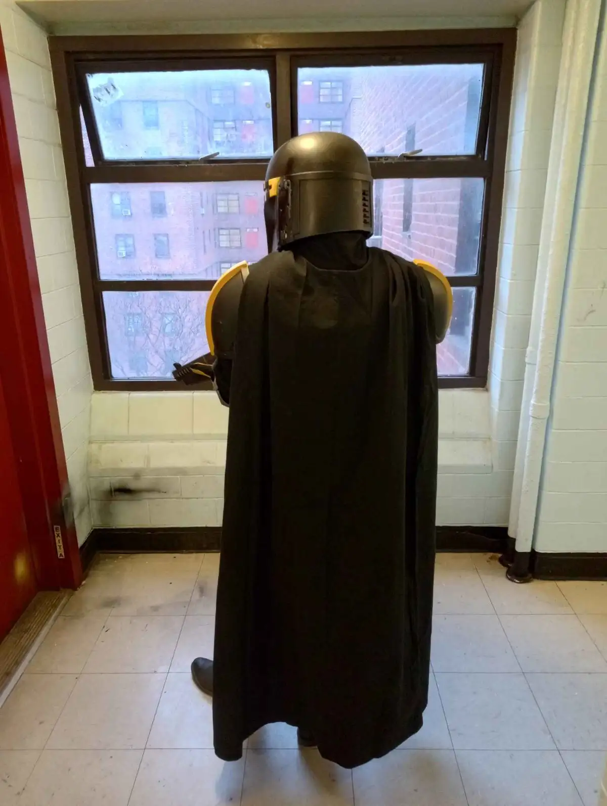A person in a full Mandalorian armor costume, viewed from behind, stands in a plain hallway looking out a window at brick…