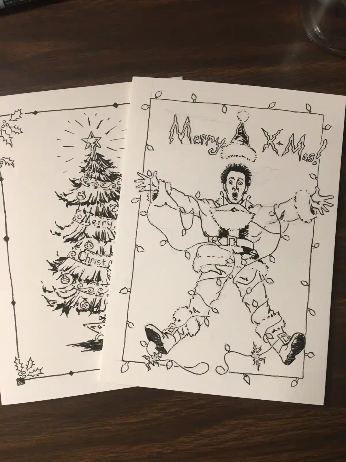 Two hand-drawn Christmas cards lie on a wooden surface. The left card shows a decorated Christmas tree with 'Merry…