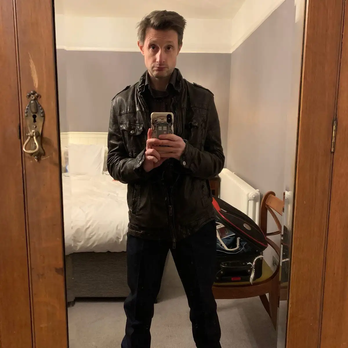 A man in a black leather jacket takes a mirror selfie in a hotel room, holding a phone with a Doctor Who TARDIS case. Behind…