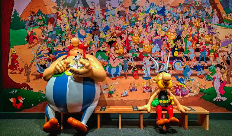 Two large statues of Asterix and Obelix sit on a bench in front of a massive, colorful mural depicting hundreds of Asterix…