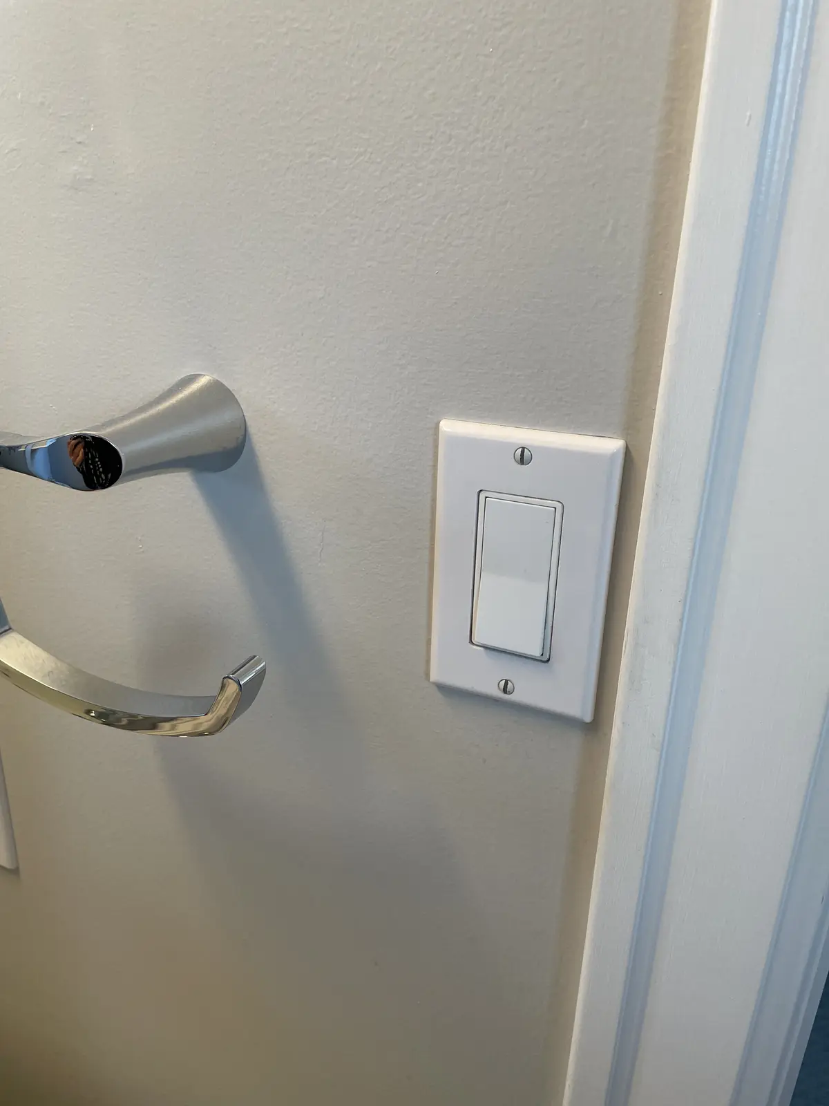 A close-up photo shows a white light switch mounted on a light-colored wall next to a shiny chrome door handle. The switch…