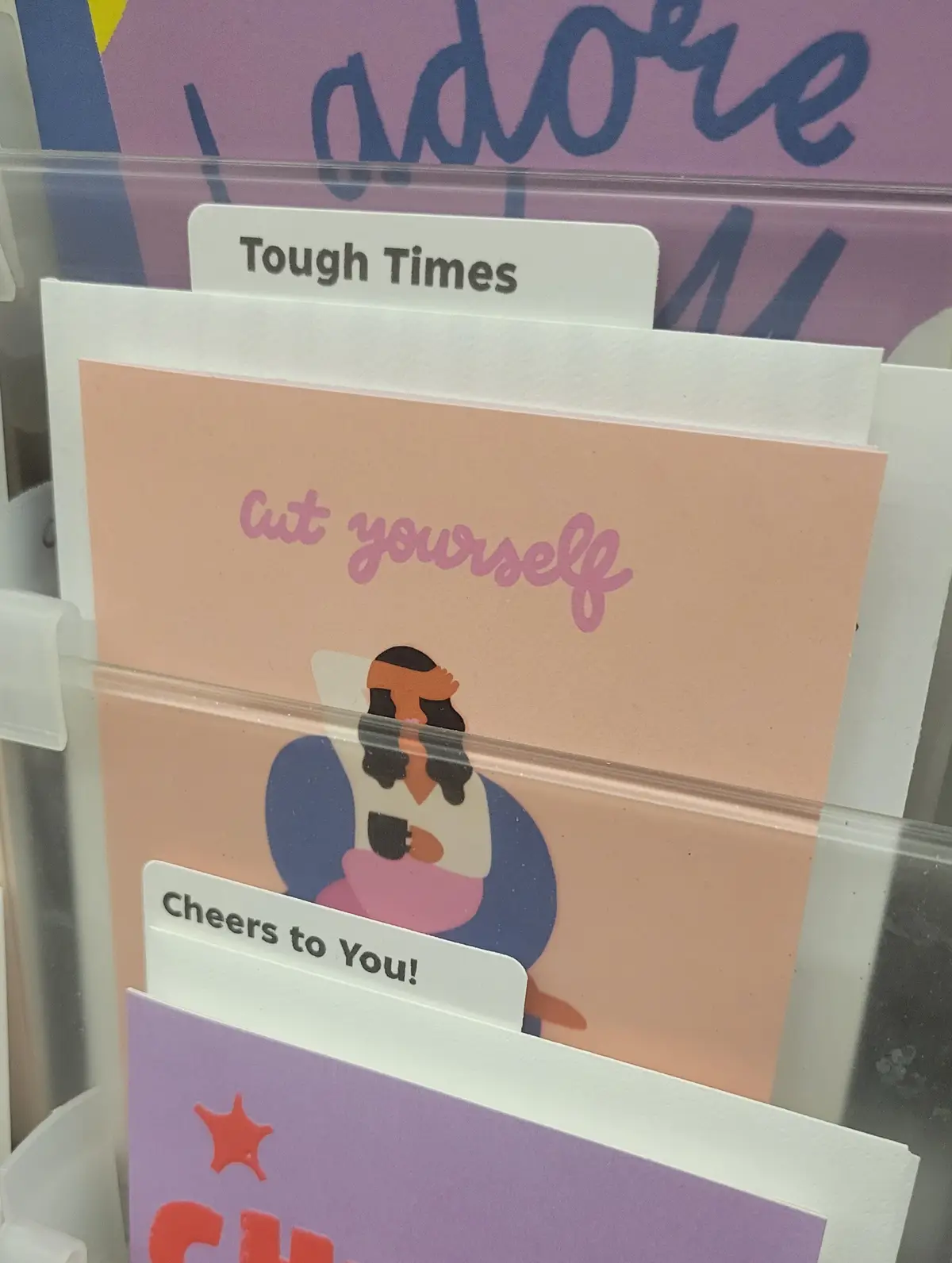 A close-up of greeting cards in a display rack, with a visible tab labeled 'Tough Times' above a card that reads 'Cut…