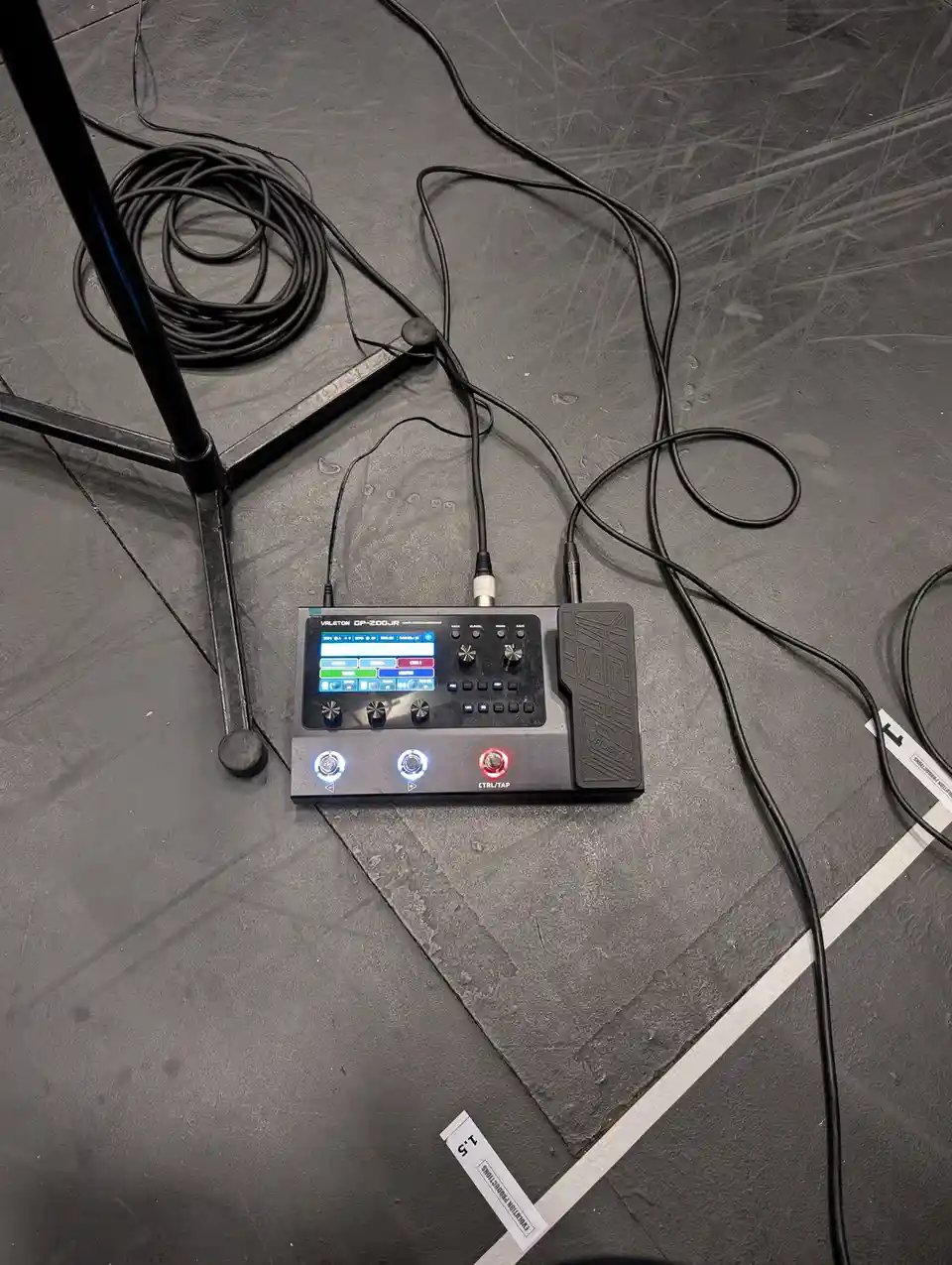 A black Valeton GP-200JR multi-effects processor sits on a scratched gray stage floor, connected by cables to a microphone…