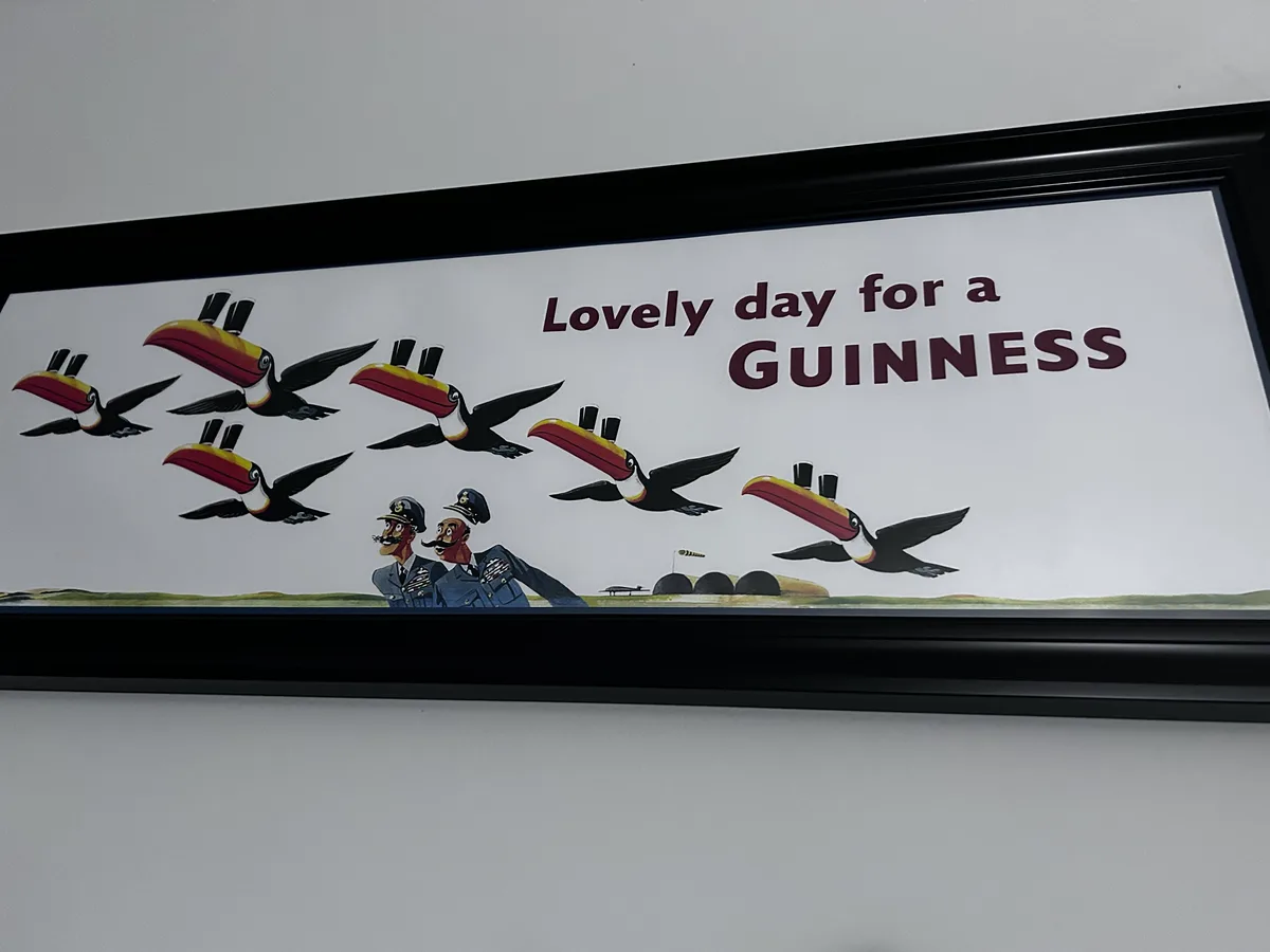 A framed vintage advertisement shows two cartoon men in naval uniforms looking up at a flock of toucans flying overhead…