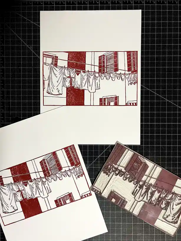 Three prints of a red and white linocut-style artwork depicting laundry hanging on a clothesline between buildings are…