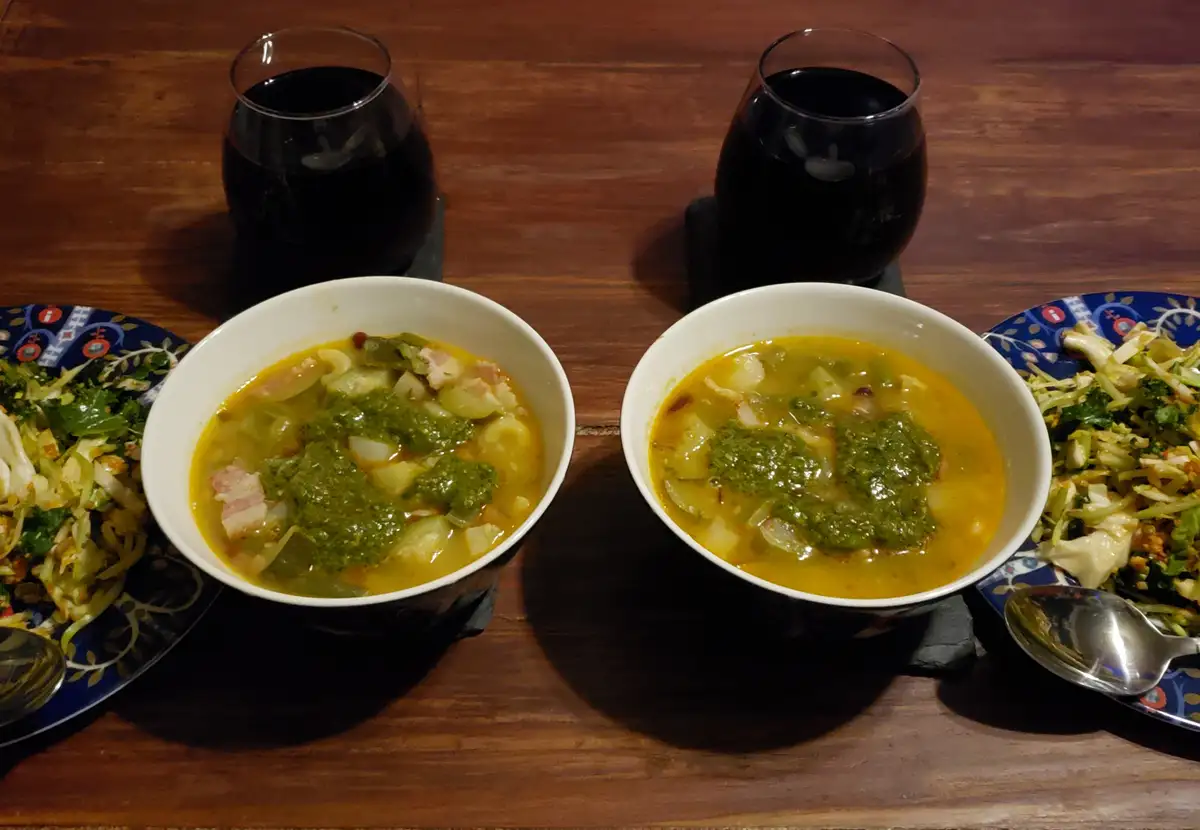 Two identical white bowls of yellowish soup topped with green pesto sit side-by-side on a wooden table, flanked by two…