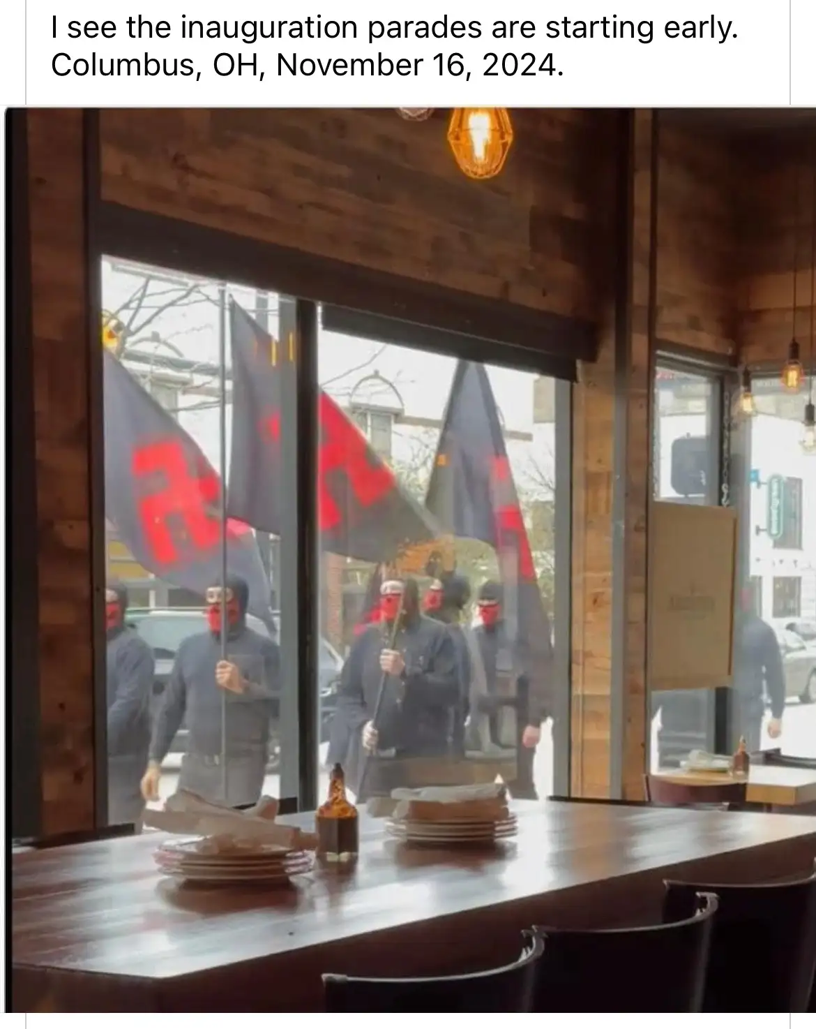 A photo taken from inside a restaurant looking out a window shows a group of people wearing red face coverings and dark…