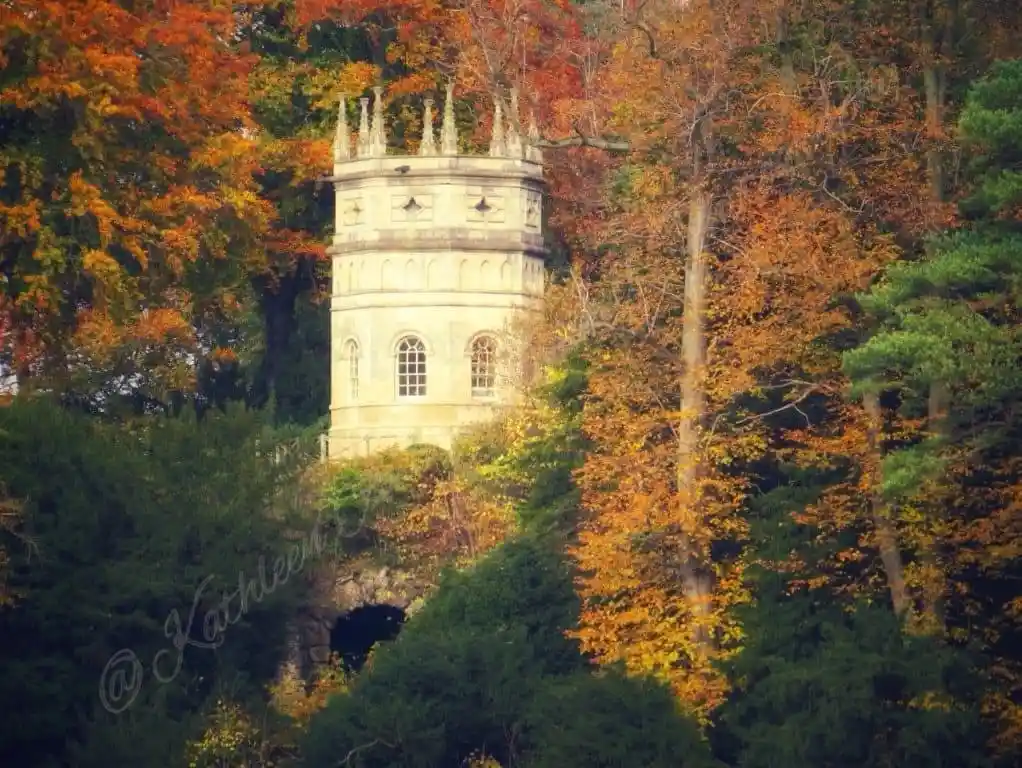 A stone tower with crenellated top sits nestled among dense trees displaying vibrant autumn foliage in shades of orange…