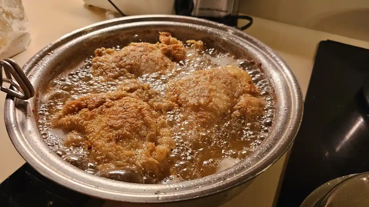 A close-up photo shows several breaded chicken pieces frying in bubbling oil inside a metal pot on a stovetop. The chicken…