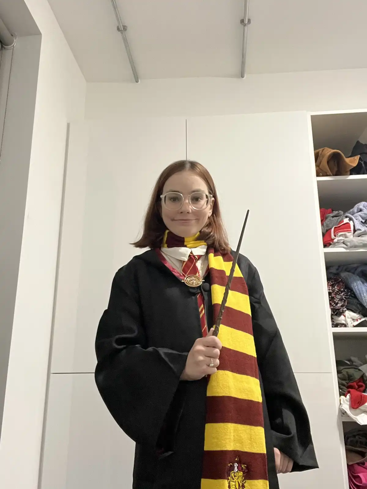 A young woman with glasses stands in a room wearing a black robe, a red-and-yellow striped Gryffindor scarf, a white…
