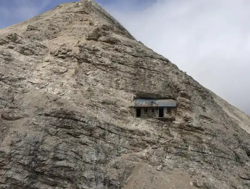 A small, weathered stone building with a metal roof is embedded into the steep, rocky face of a mountain under a cloudy sky…