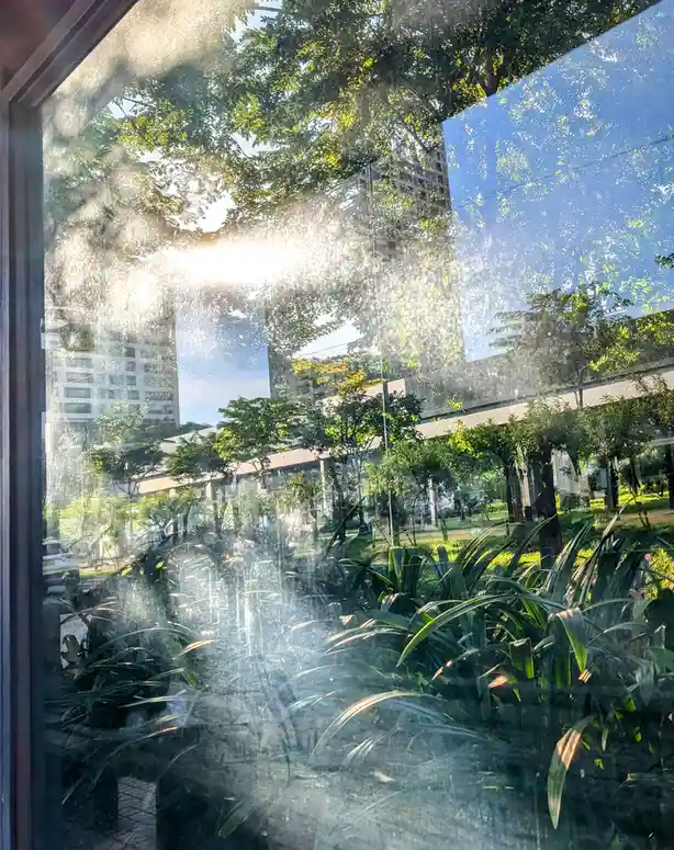 A view from inside a building looking out through a window covered in condensation or water droplets, with bright sunlight…