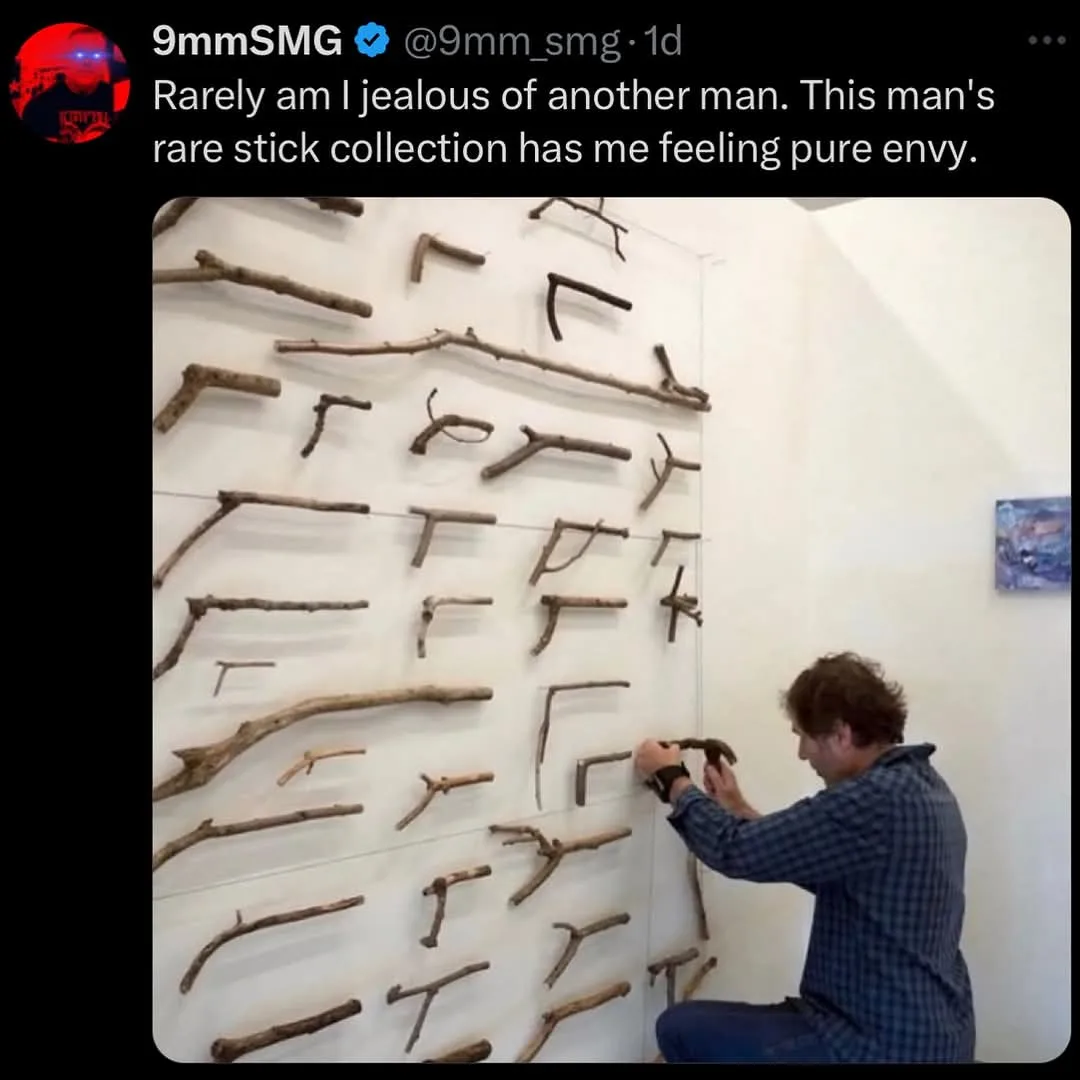 A man in a blue plaid shirt kneels while arranging various natural sticks and twigs on a white wall, which are mounted in a…