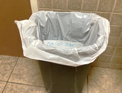 A dark-colored trash can lined with a white plastic bag sits on a tiled floor against a tiled wall. A small piece of paper…