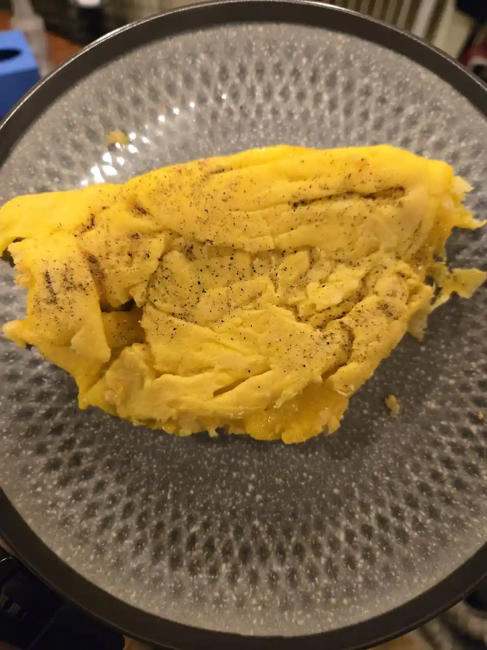 A close-up photo shows a yellow, folded omelette on a speckled gray plate, sprinkled with black pepper. The omelette's shape…