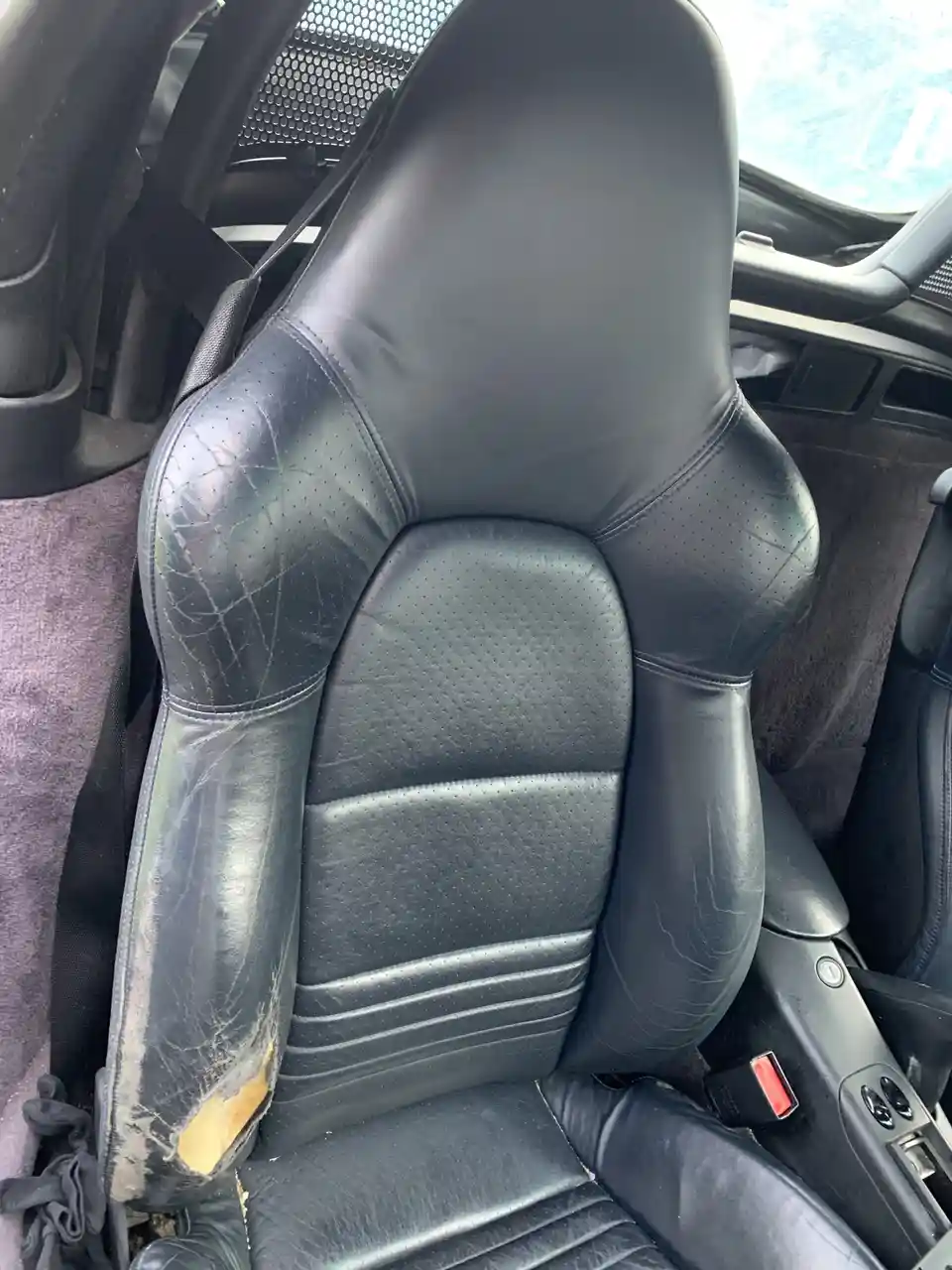 a close-up photo of a heavily worn black leather car seat with visible cracks, peeling material, and a torn section on the…