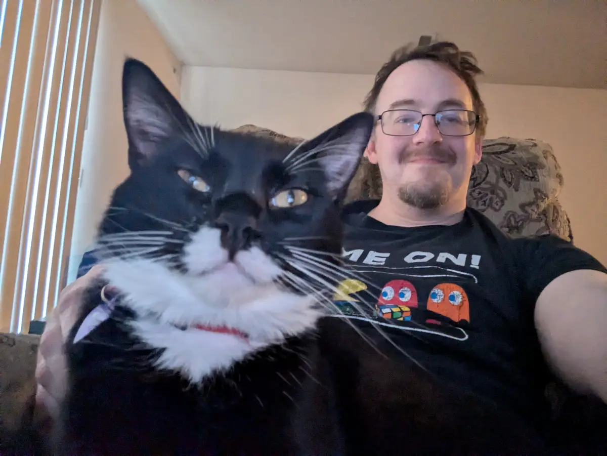 A man wearing glasses and a black t-shirt with Pac-Man ghosts and the text 'ME ON!' takes a selfie while holding a large…