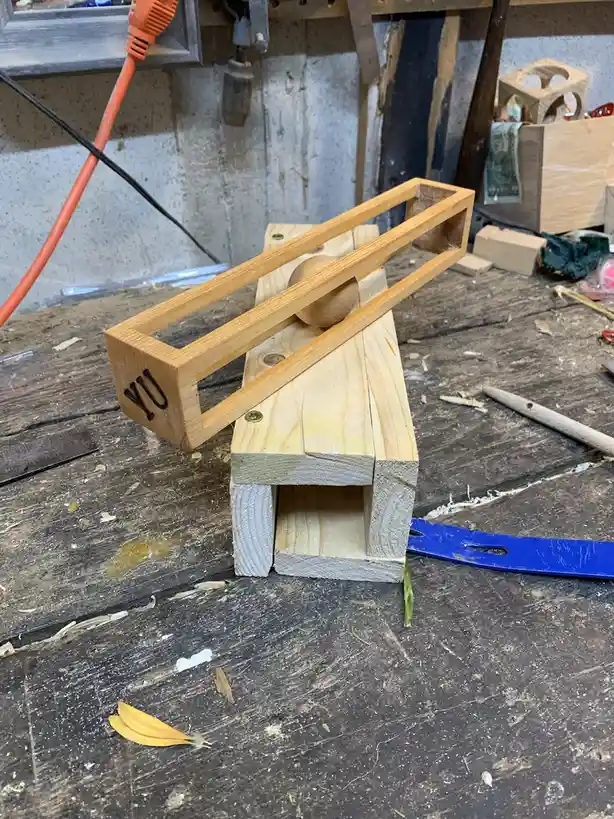 A wooden contraption on a cluttered workbench features a ball resting in a channel, with the letters 'YU' burned into one…