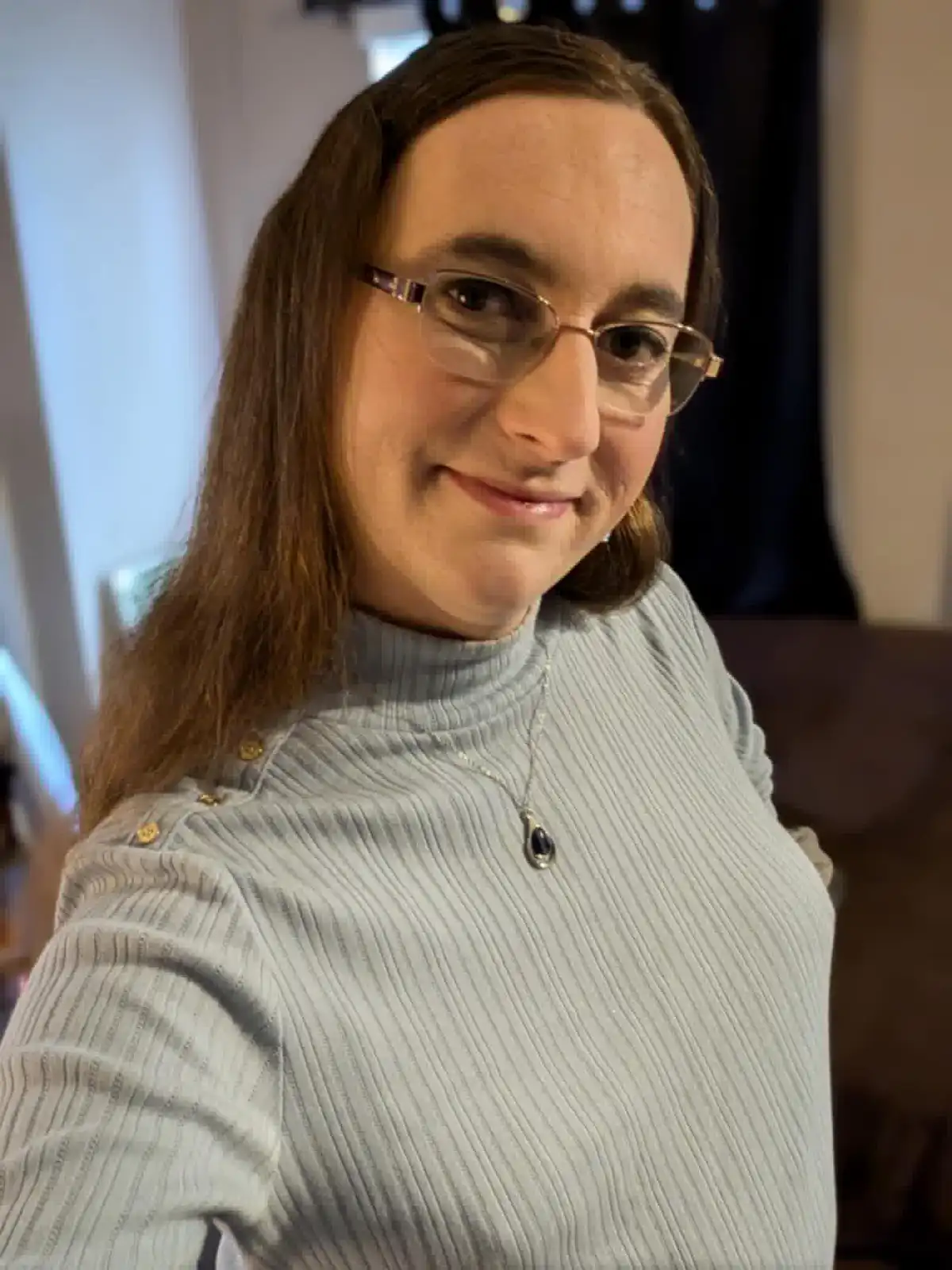 A person with long brown hair and glasses, wearing a light blue ribbed turtleneck with gold buttons on the shoulders and a…