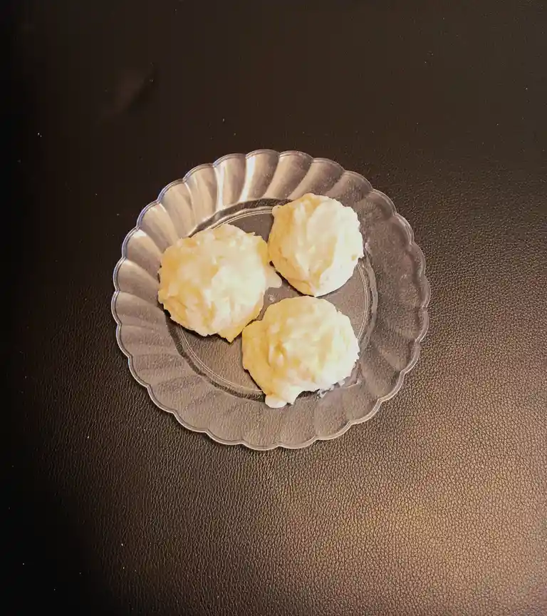 Three pale, lumpy, white-frosted cookies sit on a clear, scalloped glass plate atop a dark, textured surface. The overlaid…