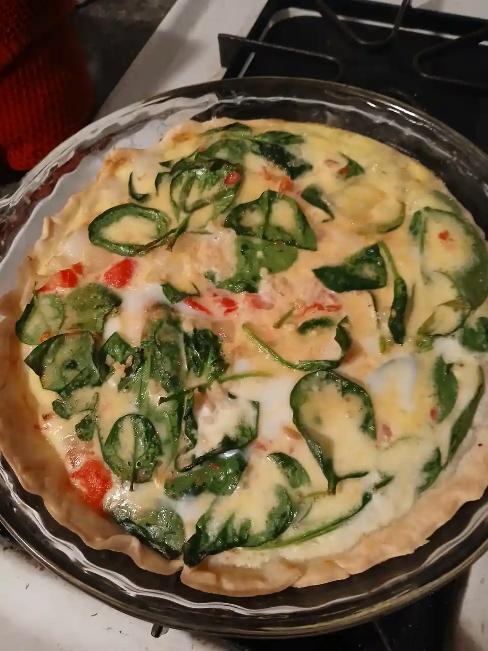 A close-up photo shows a round, unbaked quiche in a glass pie dish, featuring a pale crust and a filling of eggs, spinach…