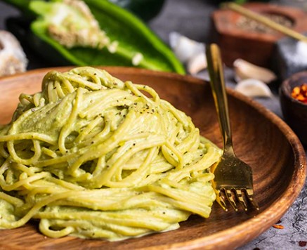 a wooden bowl holds a mound of creamy green pasta, likely pesto, with a gold fork resting beside it. The background includes…
