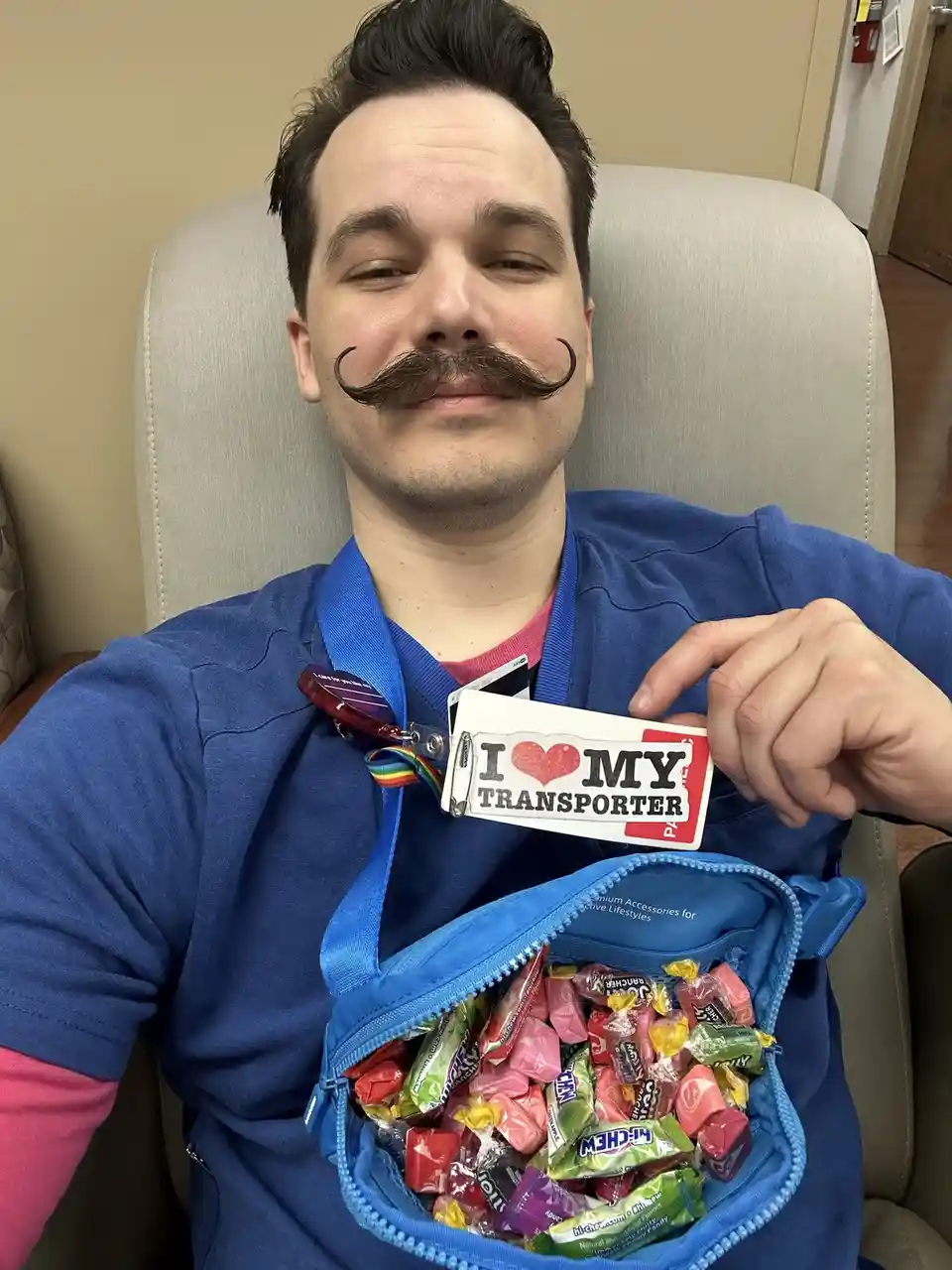 A man in blue scrubs with a fake handlebar mustache sits in a chair, holding up a badge that reads 'I ❤️ MY TRANSPORTER'…
