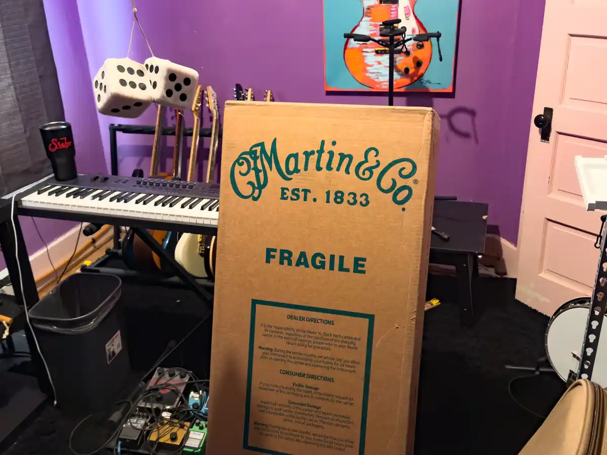 A large cardboard box labeled 'Martin & Co. EST. 1833' and 'FRAGILE' stands in a music room filled with guitars, a keyboard…