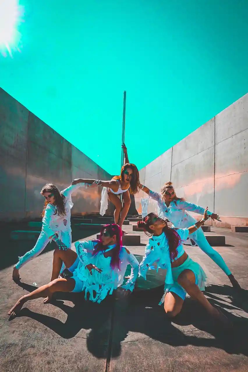 Five women in ripped, light-blue outfits pose dramatically in a sunlit concrete courtyard with a tall pole, their faces…
