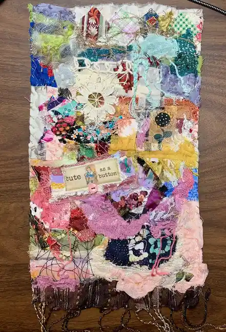 A colorful, textured patchwork textile artwork made from various fabrics, lace, and threads, resting on a wooden surface. A…