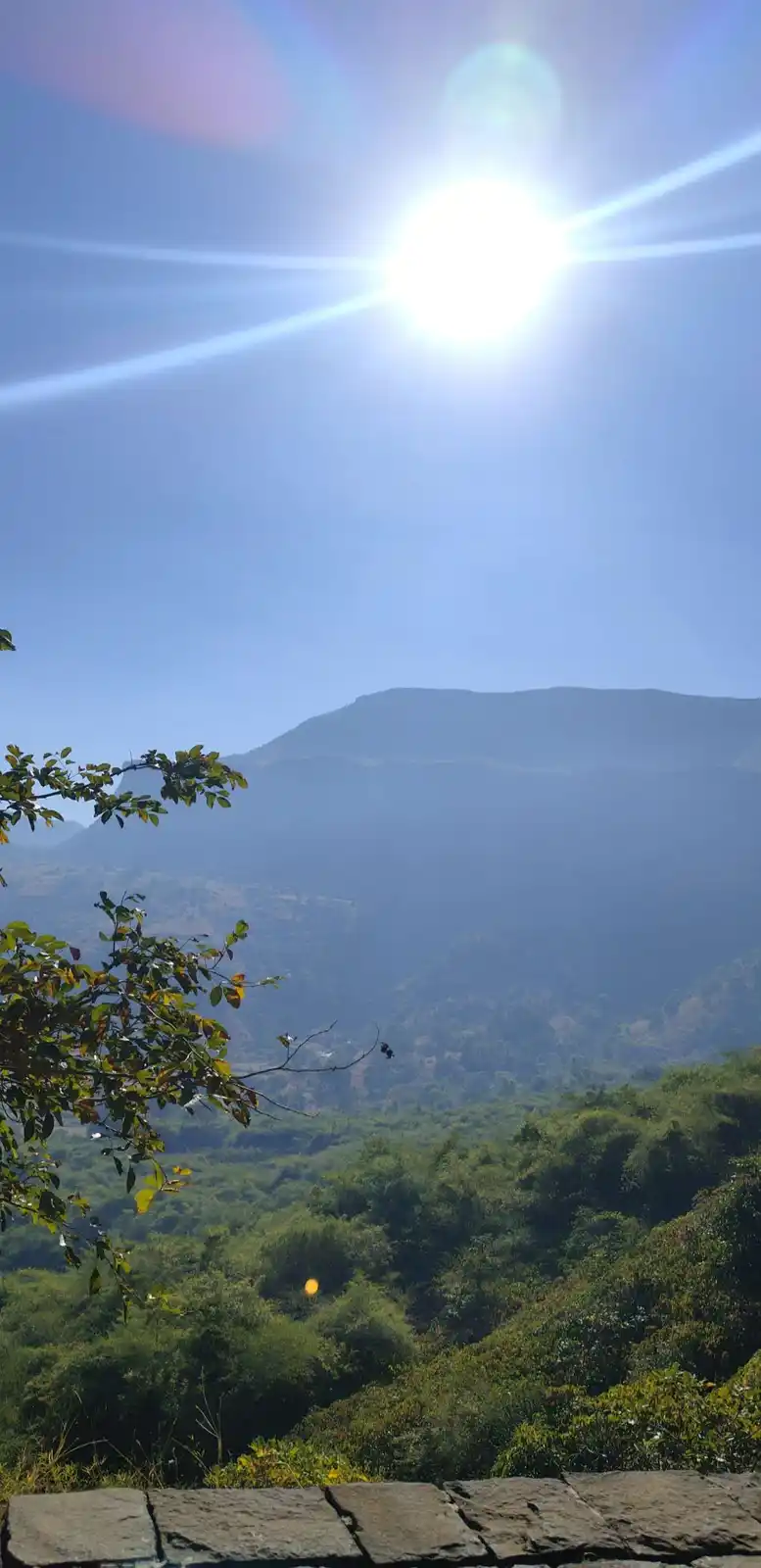 A bright sun shines overhead in a clear blue sky above a mountain range with lush green forests below. A stone wall is…