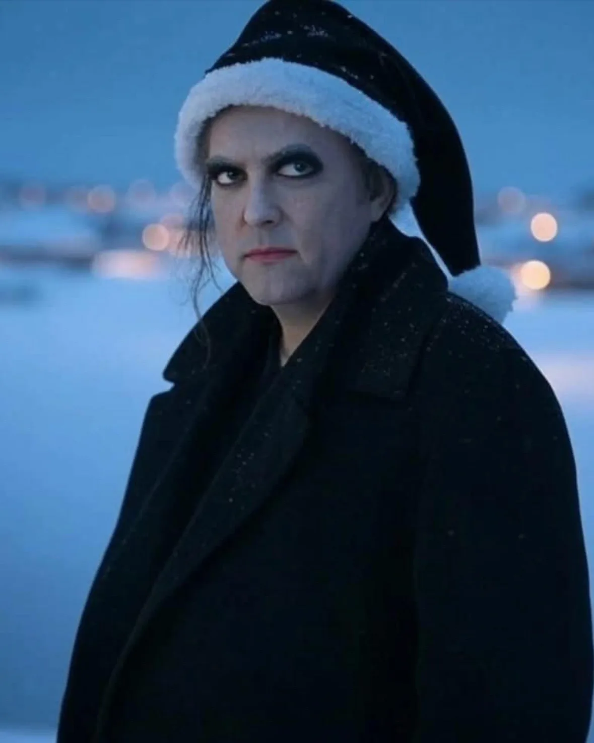 a man with heavy dark eye makeup and pale skin wears a black santa hat with white trim and a black coat while standing in a…