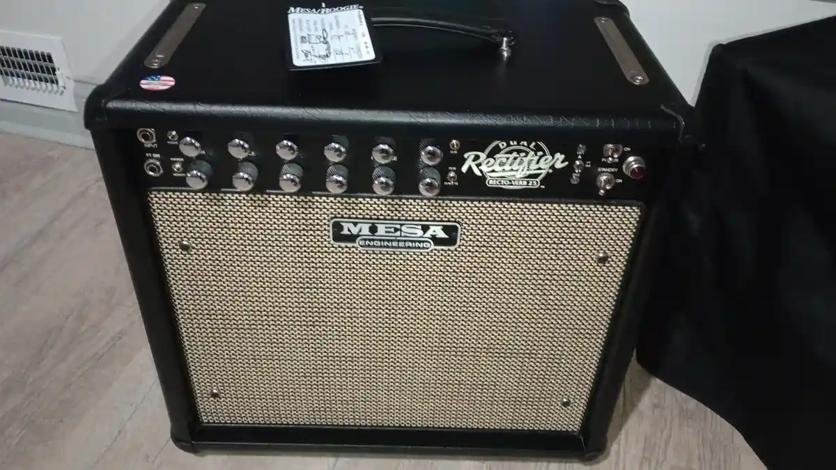 A black Mesa Boogie Dual Rectifier guitar amplifier head sits on a wooden floor, featuring a beige grille cloth, numerous…